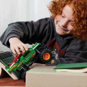 Joyful child with red hair playing with a green monster truck on a cozy carpet.