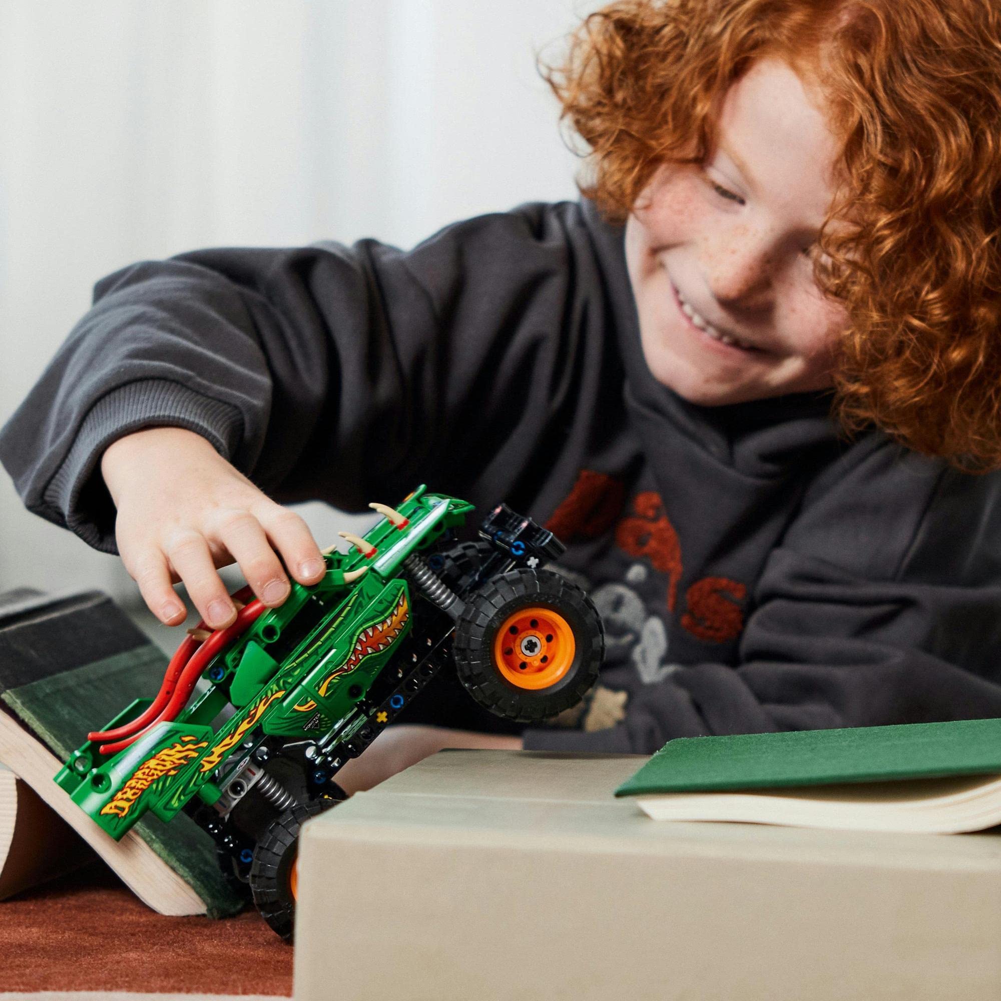 Joyful child with red hair playing with a green monster truck on a cozy carpet.