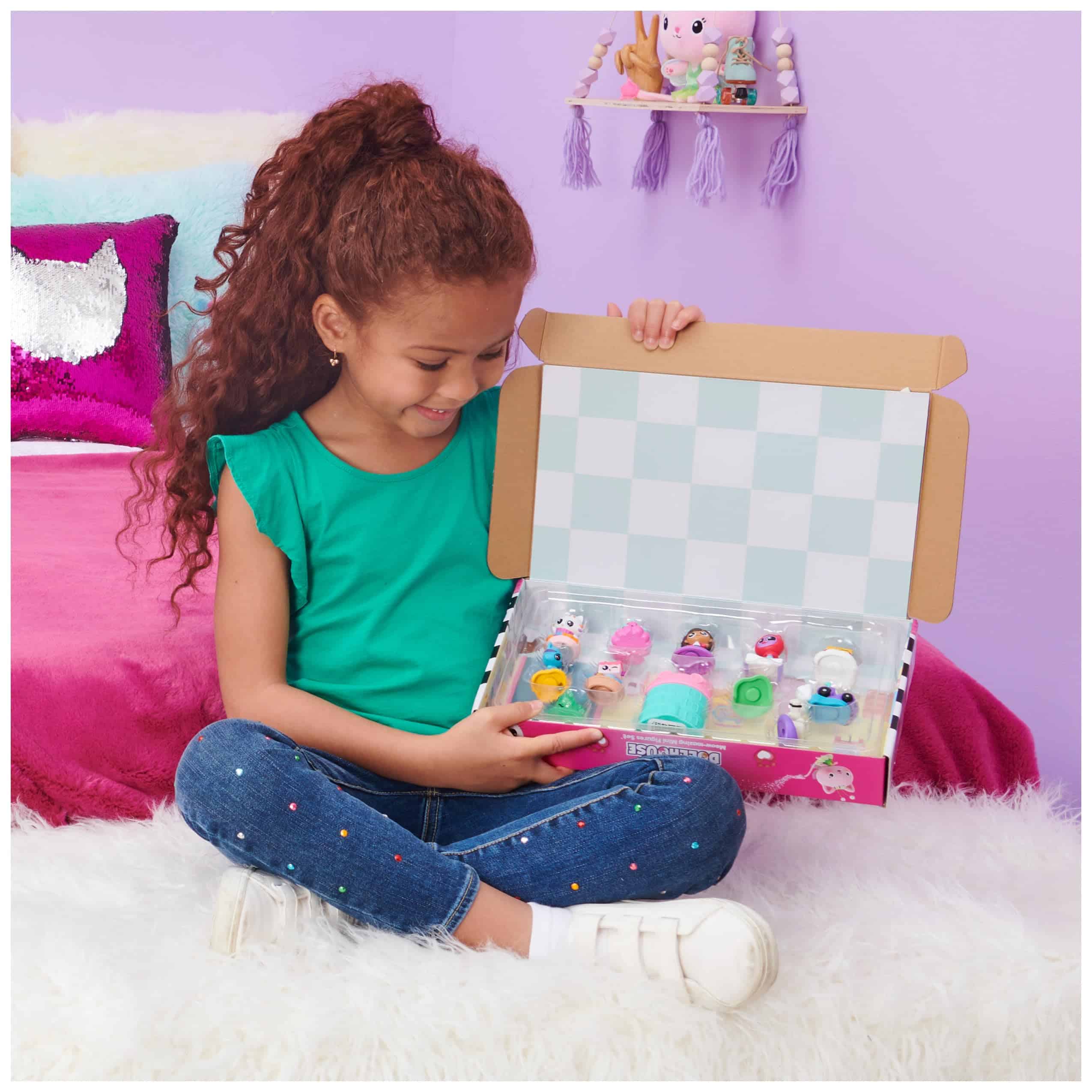 Young girl joyfully unboxing colorful toys on a plush pink bed against a purple wall.