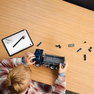 Person assembling a gray model truck with digital tablet guidance on a wooden table.