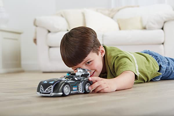 Boy plays with a glossy race car on a light wooden floor in a cozy living room.